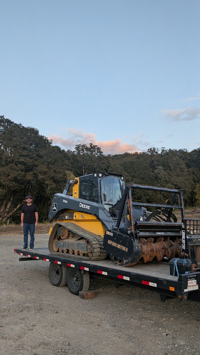 Owner standing next to the John Deere track loader and mulching head loaded on a flatbed trailer at dusk